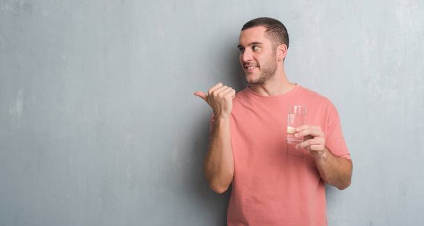 Kaukasische jongeman over grijs grunge muur drinken van een glas water aan te wijzen en te tonen met de duim aan de kant met een blij gezicht lachend - Foto, afbeelding