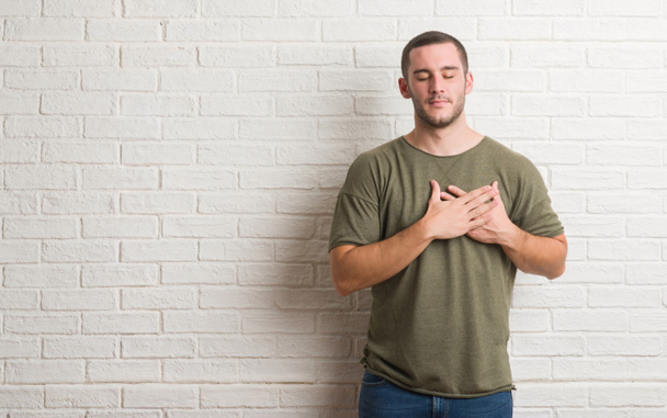 Young caucasian man standing over white brick wall smiling with hands on chest with closed eyes and grateful gesture on face. Health concept. - Photo, Image