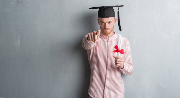 junger rothaariger Mann über grauer Grunge-Wand mit Diplommütze, der mit dem Finger in die Kamera und zu dir zeigt, Handzeichen, positive und selbstbewusste Geste von vorne - Foto, Bild