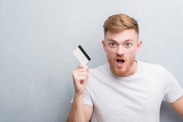 Jonge roodharige man over grijs grunge muur creditcard bang in schok met een verrassing gezicht, bang en opgewonden met angst expressie te houden - Foto, afbeelding