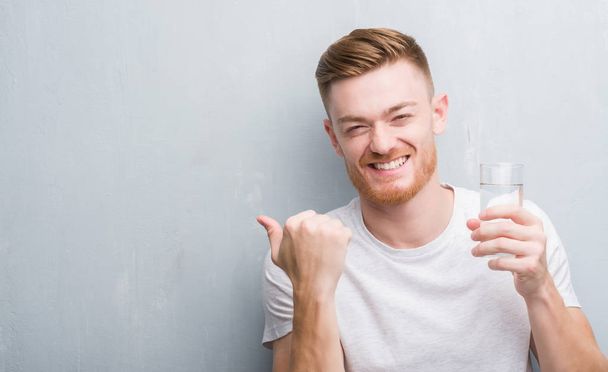 Young redhead man over grey grunge wall drinking water pointing and showing with thumb up to the side with happy face smiling - Фото, изображение