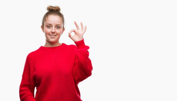 Молодая блондинка с булочкой и красным свитером, улыбаясь позитивно, делает знак "хорошо" с рукой и пальцами. Успешное выражение
. - Фото, изображение