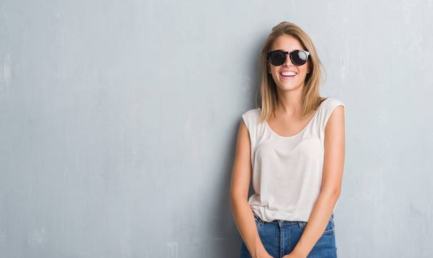 Beautiful young woman standing over grunge grey wall wearing fashion sunglasses with a happy face standing and smiling with a confident smile showing teeth - Фото, зображення
