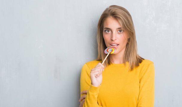 Beautiful young woman over grunge grey wall eating lollipop candy with a confident expression on smart face thinking serious - Photo, Image