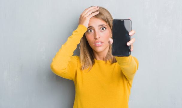 Mooie jonge vrouw over grunge grijs muur tonen smartphone scherm benadrukt met de hand op het hoofd, geschokt met schaamte en verrassing gezicht, boos en gefrustreerd. Angst en boos voor fout. - Foto, afbeelding