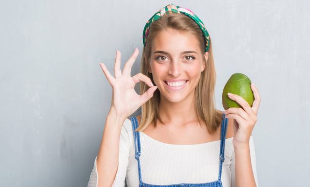 Kaunis nuori nainen yli grunge harmaa seinä tilalla tuoretta mango tekee ok merkki sormilla, erinomainen symboli
 - Valokuva, kuva