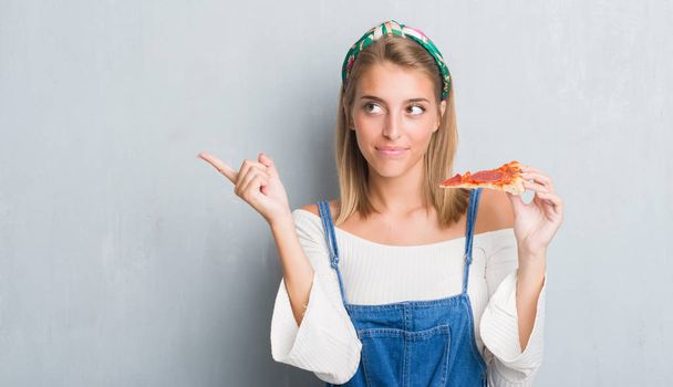 Mulher bonita sobre parede cinza grunge comendo fatia de pizza pepperoni muito feliz apontando com a mão e o dedo para o lado
 - Foto, Imagem