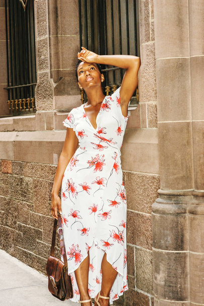 Mujer afroamericana viajando, trabajando en Nueva York, vistiendo un vestido largo con dibujos blancos, llevando a mano un bolso de cuero marrón, mano descansando en la frente, de pie por la pared en la calle, mirando hacia arriba, triste, cansado
 - Foto, imagen