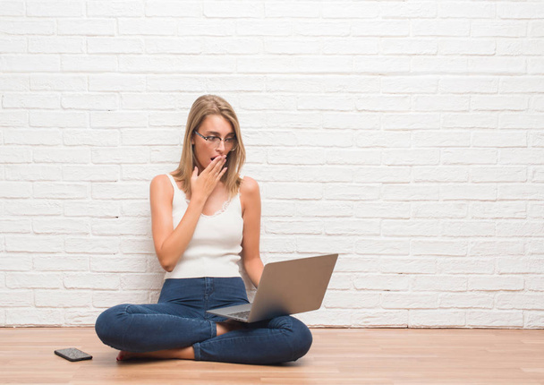 Mulher bonita sentada no chão trabalhando usando laptop em casa cobrir a boca com a mão chocada com vergonha de erro, expressão de medo, assustado em silêncio, conceito secreto
 - Foto, Imagem