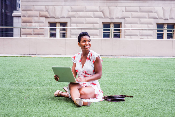 African American College Student studying in New York, with short afro hair, wearing long dress, sandal high heels, crossing legs, sitting on green lawn on campus, working on laptop computer, smiling - Photo, Image