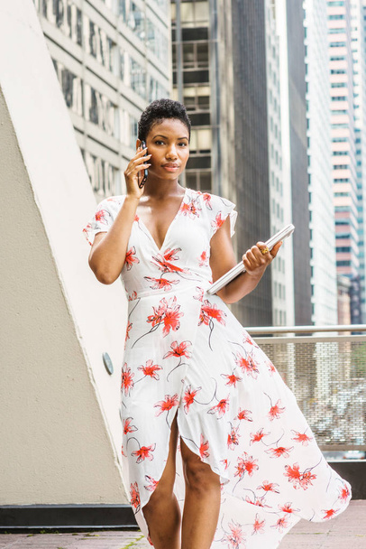 Young African American Businesswoman with short afro hair, wearing long dress, holding laptop computer, talking on cell phone, walking on balcony in New York. Street with hight buildings on background - Photo, Image