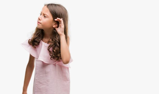 Brunetka hispánský dívka, která nosí růžové šaty s úsměvem s rukou přes ucho naslouchá slyšení zvěsti nebo drby. Hluchota koncept. - Fotografie, Obrázek