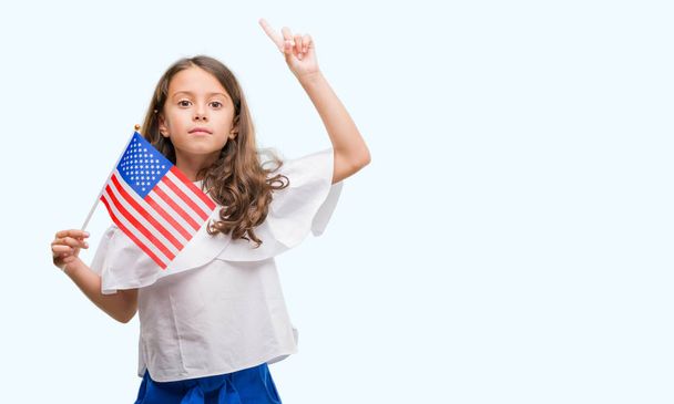 Chica hispana morena sosteniendo bandera de Estados Unidos de América sorprendida con una idea o pregunta apuntando con el dedo con la cara feliz, número uno
 - Foto, imagen