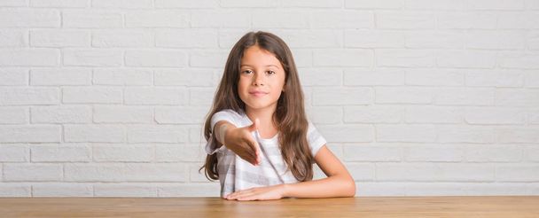 Młode dziecko hiszpanin siedzi na stole w domu uśmiechnięty oferuje przyjazny uścisk dłoni jako pozdrowienie i przyjazna. Udanego biznesu. - Zdjęcie, obraz