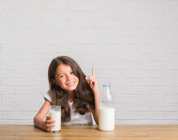 幸せそうな顔でのアイデアや質問ポインティング指に驚いてミルクのガラスを飲むテーブルに座って若いヒスパニックの子供がナンバーワンします。 - 写真・画像