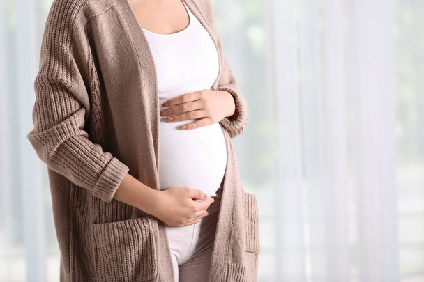 Pregnant woman standing near window at home, closeup - Foto, afbeelding