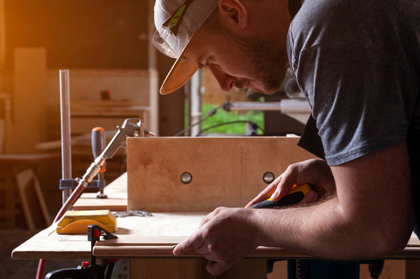 Een bouwer van de jonge brunette man dragen groene zwembrillen, een grijs T-shirt en een baard, een houten product behandelen met een beitel in de werkplaats, close-up - Foto, afbeelding