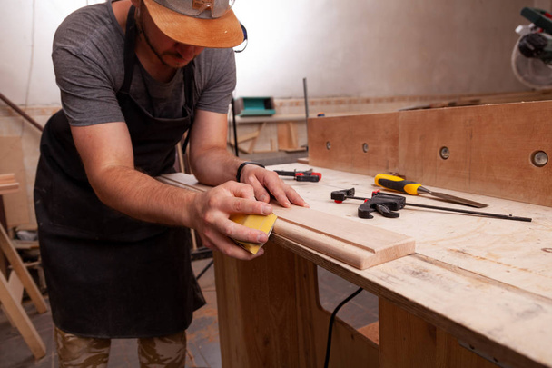 Primer plano de Un trabajador con gorra y camisa pule el bloque de madera con papel de lija antes de pintar en el taller
 - Foto, imagen