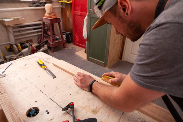 Primer plano de Un trabajador con gorra y camisa pule el bloque de madera con papel de lija antes de pintar en el taller
 - Foto, imagen