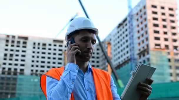 Adult engineer or architect uses a tablet in operation. Writes a message or checks a drawing. Against background is building. Builders are building a modern residential building of glass and concrete. - Video