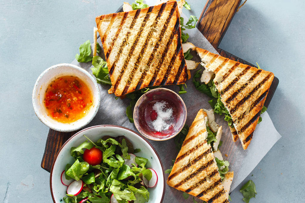 Grilled sandwich with chicken, arugula on wooden cutting board with salad, sauce and pickled cucumbers top view on blue background. Nutritious and healthy breakfast - Φωτογραφία, εικόνα