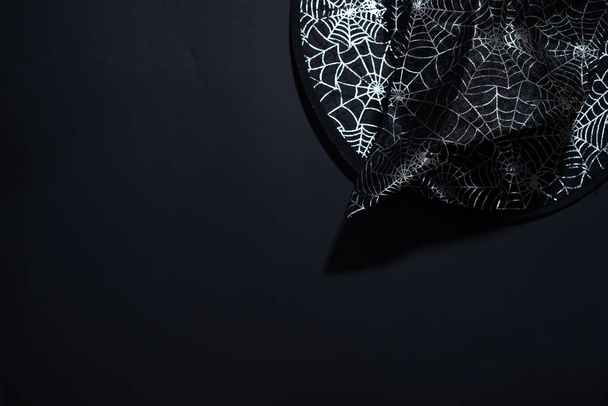 Black witch hat with a pattern of cobwebs on black background, top view. Halloween background with copy space - Photo, Image