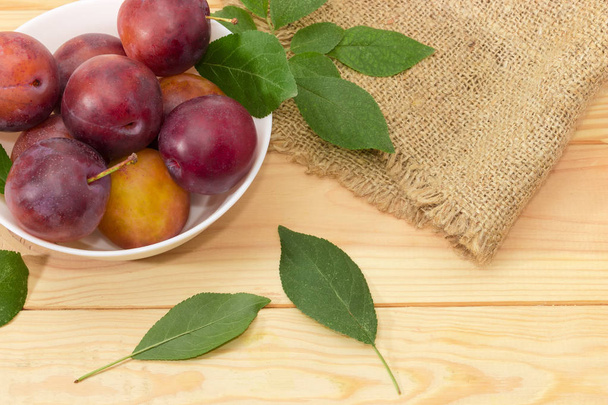 Fragment of the white bowl with ripe purple plums and leaves of plum tree on a light colored wooden surface and burlap - Foto, Imagen