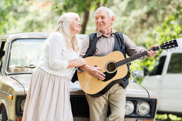 старша жінка обіймає чоловіка, який грає на гітарі проти ретро-автомобіля
 - Фото, зображення