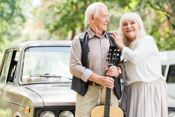 senior woman embracing man with guitar against beige vintage car  - Foto, imagen