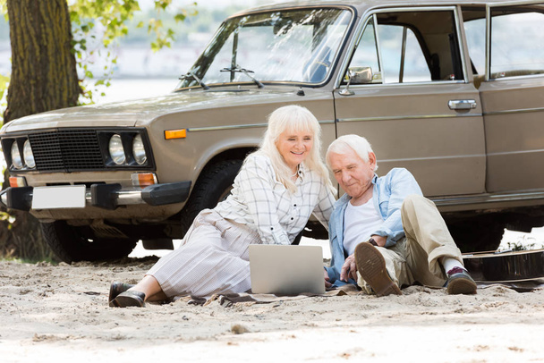 Happy senior couple sitting on sand and using laptop near beige vintage car - Photo, image