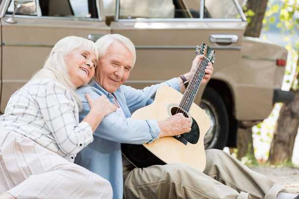 mujer mayor sentada en la arena con el hombre tocando la guitarra contra el coche vintage
  - Foto, Imagen