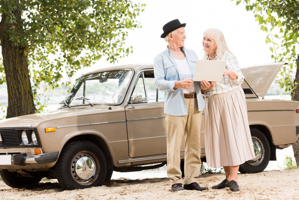 beautiful senior couple using laptop and looking at each other against beige vintage car - Fotó, kép