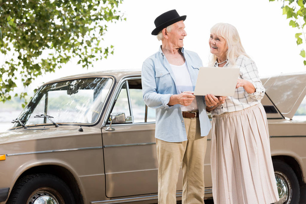 Happy senior couple using laptop and looking at each other against beige retro car - Zdjęcie, obraz