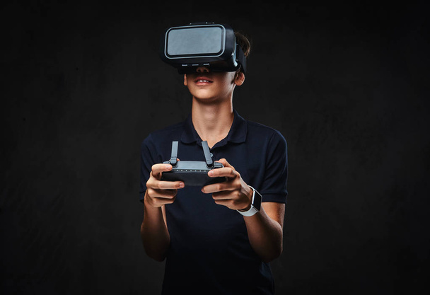 Adolescente joven vestida con una camiseta negra lleva gafas de realidad virtual y controla el quadcopter usando el control remoto. Aislado sobre un fondo oscuro
. - Foto, Imagen