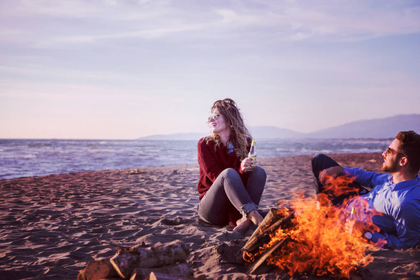 Young Couple Relaxing By The Fire, Drinking A Beer Or A Drink From The Bottle on the beach at autumn day filter - Фото, зображення