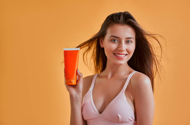 Hello, summer! Attractive young woman in swimwear isolated on orange background. Happy girl in pink sunglasses is posing with sunscreen. Body care concept. - Фото, зображення