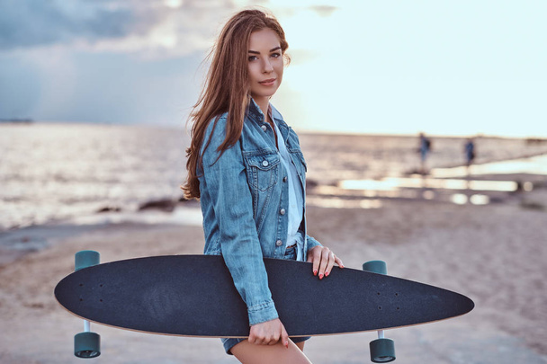 Αισθησιακό μελαχρινή κοπέλα ντυμένος με σακάκι τζιν σορτς και κατέχει skateboard ενώ στέκεται στην παραλία σε συννεφιά κατά το ηλιοβασίλεμα. - Φωτογραφία, εικόνα