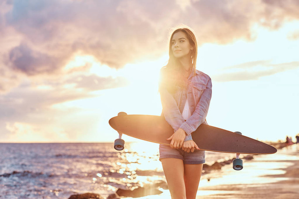 Sensuelle fille brune vêtue de short en denim et veste tient skateboard tout en se tenant sur la plage par temps nuageux lors du coucher de soleil lumineux
. - Photo, image
