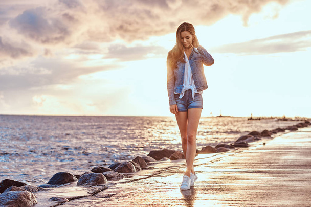 Full body portrait of a charming girl dressed in denim shorts and jacket walks on the beach in cloudy weather during bright sunset. - Photo, Image
