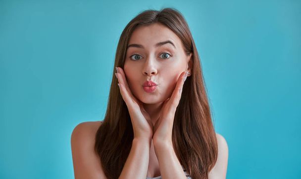 Attractive young woman isolated on blue background. Portrait of happy girl is blowing air kiss. - Zdjęcie, obraz