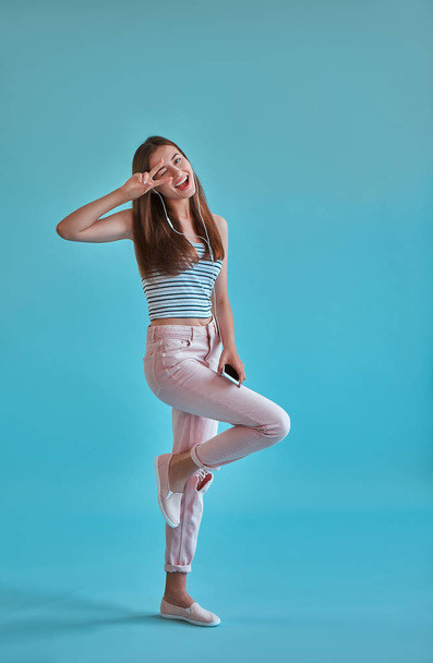 Attractive young woman isolated on blue background. Happy girl is posing with smart phone and earphones. Dancing, having fun while listening to music and showing V-sign. - Foto, afbeelding