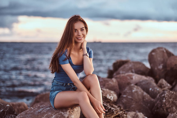 Porträt eines glücklichen, charmanten Mädchens, das bei Sonnenuntergang auf Felsen vor einem bewölkten Himmel sitzt. - Foto, Bild
