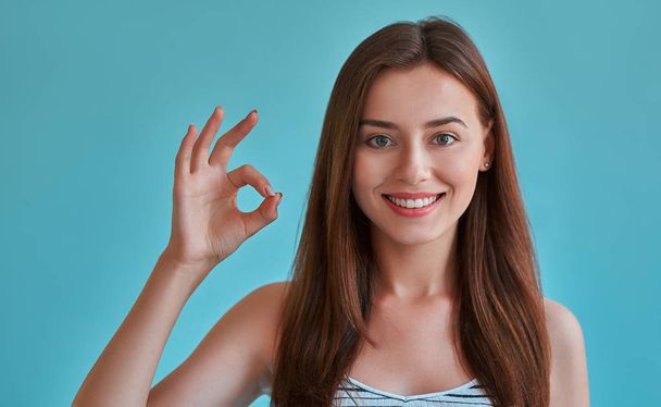 Attractive young woman isolated on blue background. Portrait of happy girl posing, smiling and showing OK sign. - Photo, Image