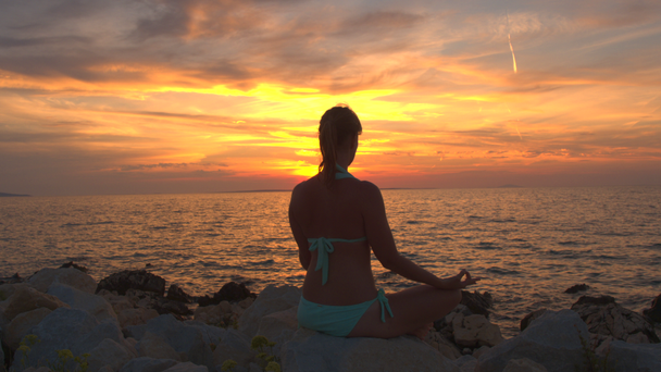 CERRAR: Chica tranquila sentada en la roca por el mar ondulante, meditando en la pose de loto yoga, concentrándose, observando la respiración y relajando la mente, el alma y el cuerpo bajo el sol ardiente brillante y el cielo poniente dorado
 - Imágenes, Vídeo