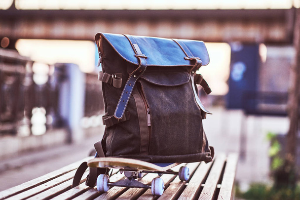 Close-up photo of a backpack lying on a skateboard that stands on a bench on the embankment during a beautiful sunset. - Fotó, kép