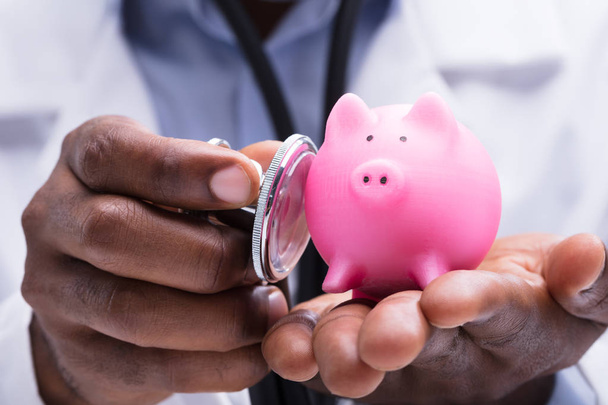 Close-up Of A Doctor Examining Pink Miniature Piggybank In His Hand With Stethoscope - Foto, immagini