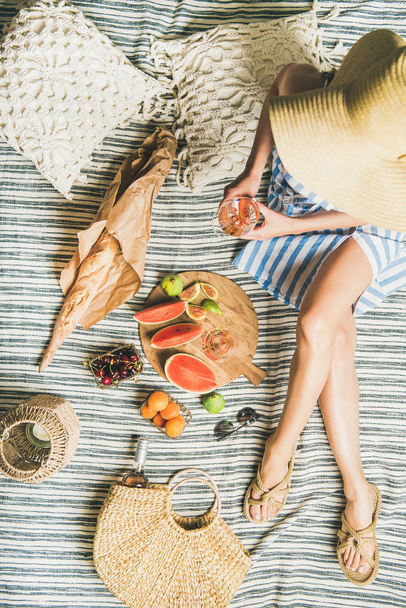 Summer picnic setting. Woman in linen striped dress and straw sunhat sitting with glass of rose wine in hand, fresh fruit on board and baguette on blanket - Foto, Imagen