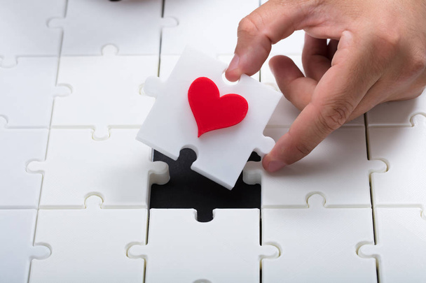 Person's hand placing last piece with red heart into white jigsaw puzzle - Foto, Imagen