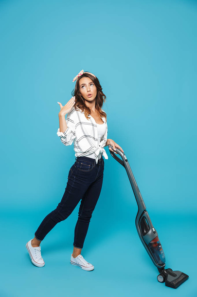 Full length portrait of tired young woman 20s housekeeping and vacuuming floor isolated over blue background - Foto, afbeelding