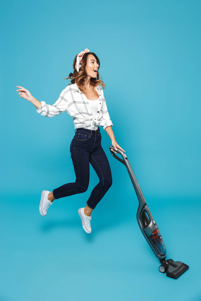 Full length photo of funny housewife 20s listening to music via wireless headphones and jumping with vacuum cleaner isolated over blue background - Foto, imagen
